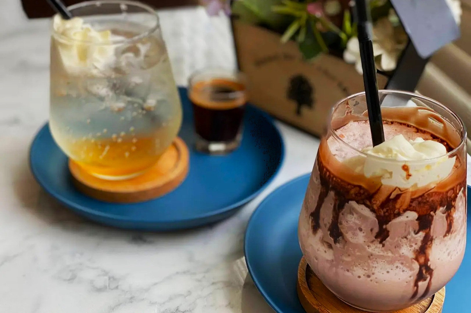 Two drinks on blue plates: a fizzy drink with cream and an iced chocolate with cream and syrup. A small espresso sits between them on a marble table.