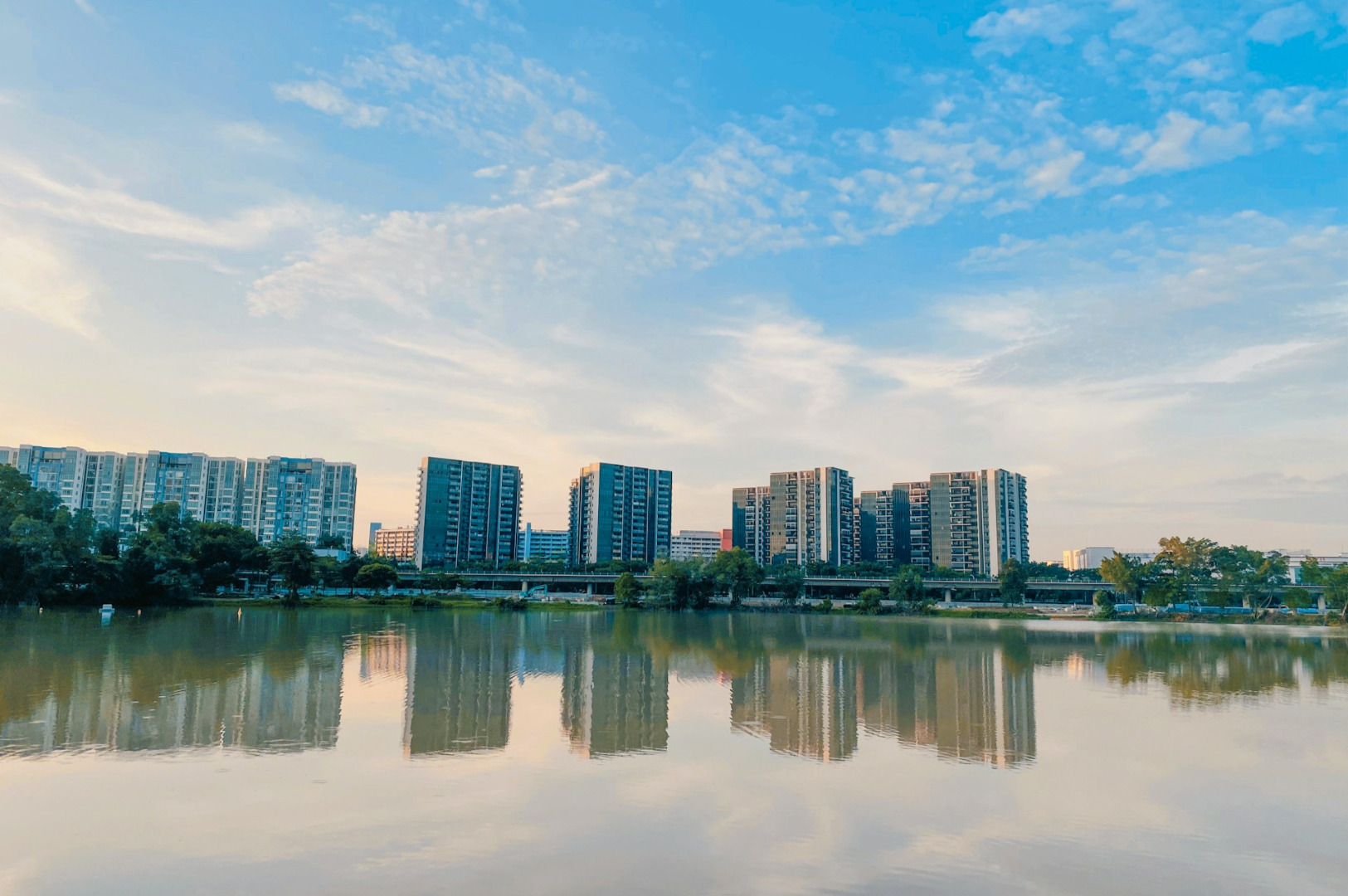 Panoramic view of high-rise buildings reflected in a calm lake under a bright blue sky with scattered clouds, creating a serene and tranquil atmosphere.