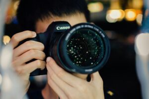 Person holding a Canon camera close to their face, focusing on the lens. Background is softly blurred with bokeh lights, creating a dreamlike ambiance.