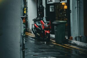 Red scooter parked in a dimly lit urban alley with wet pavement, surrounded by towering buildings and a green trash bin, creating a moody, quiet atmosphere.