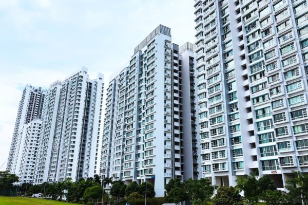 Tall, modern apartment buildings with numerous windows stand side by side under a cloudy sky, surrounded by lush green trees and a grassy area.