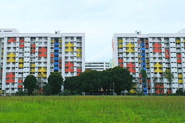 Three high-rise apartment buildings feature colorful panels of red, yellow, and blue on their facades. A vibrant green field and lush trees are in the foreground. The scene conveys a lively urban environment against a cloudy sky.