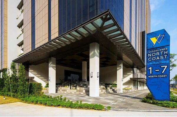 A modern building with a glass facade and entrance canopy. A large blue sign reads "Woodlands North Coast 1-7." Lush greenery lines the pathway, creating an inviting atmosphere.