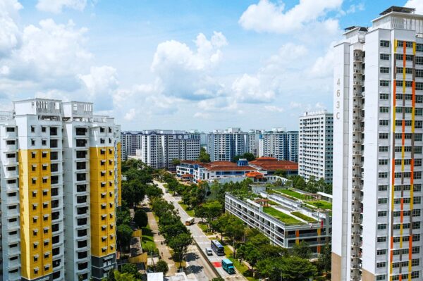 Aerial view of a residential area with tall, colorful apartment buildings under a partly cloudy sky. A road lined with trees runs through, conveying a sense of urban calm.