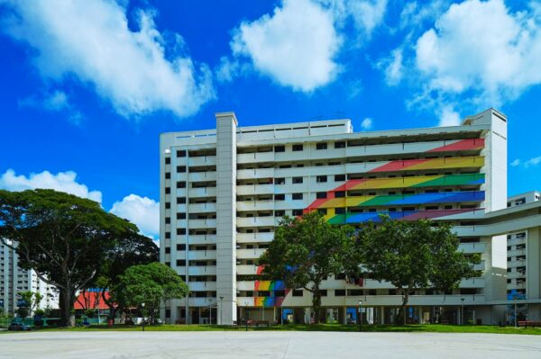 A colorful apartment building features a rainbow stripe pattern on its facade, set against a vibrant blue sky with fluffy clouds. Green trees line the front, adding a lively touch.