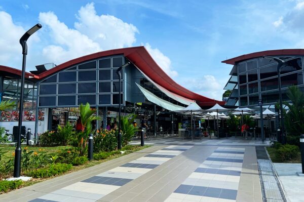 Modern building with wavy red roof and large glass windows, surrounded by lush greenery. A paved path leads to outdoor seating, under a blue sky.