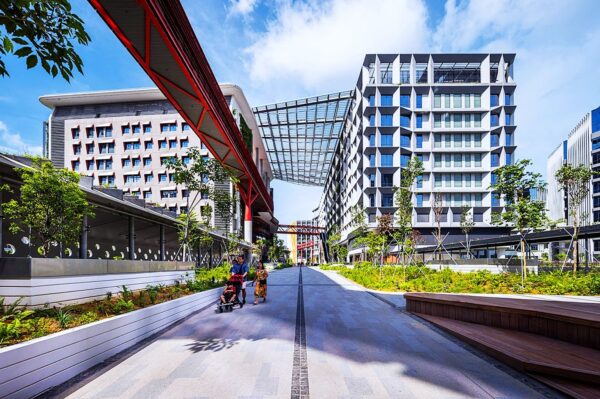 A modern, urban plaza flanked by geometric buildings under a blue sky. A red monorail track crosses overhead. People casually stroll, conveying a relaxed vibe.