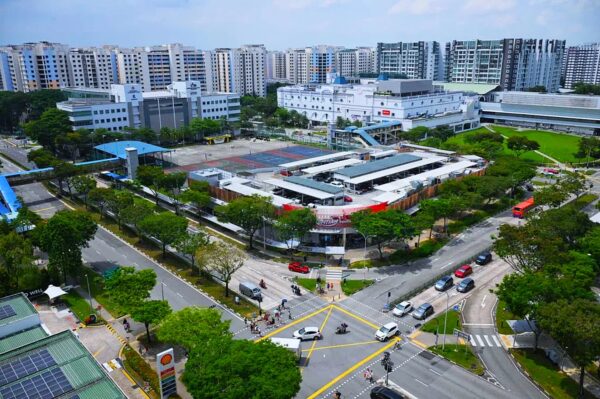 Aerial view of a busy urban intersection surrounded by trees, featuring buildings and a large white complex in the center. High-rise apartments in the background.