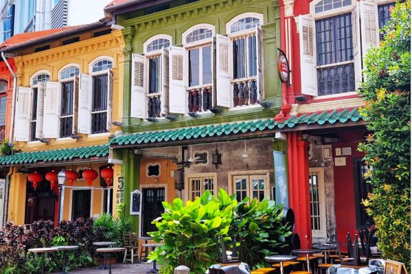 Colorful, historic buildings in warm hues of yellow, green, and red with white shutters. Lush greenery and traditional lanterns create a vibrant, inviting atmosphere.