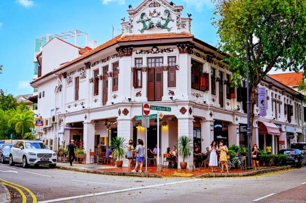 A vibrant street corner with a white heritage building featuring ornate details, red shutters, and a terracotta roof. People dine outdoors, enjoying a lively, sunny atmosphere.