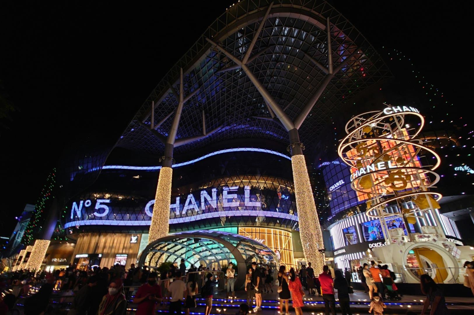 Nighttime view of a vibrant shopping mall entrance, featuring illuminated Chanel signage. Throngs of people gather under colorful lights, creating a lively atmosphere.