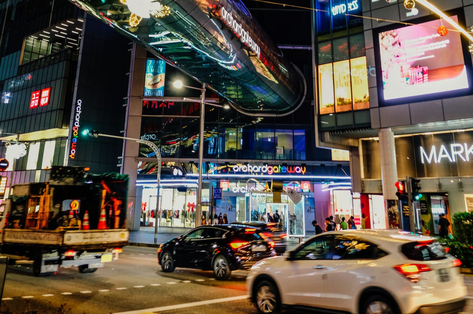 Bustling night scene at Orchard Gateway, Singapore. Bright lights, vibrant storefronts, and busy traffic create a dynamic urban atmosphere.