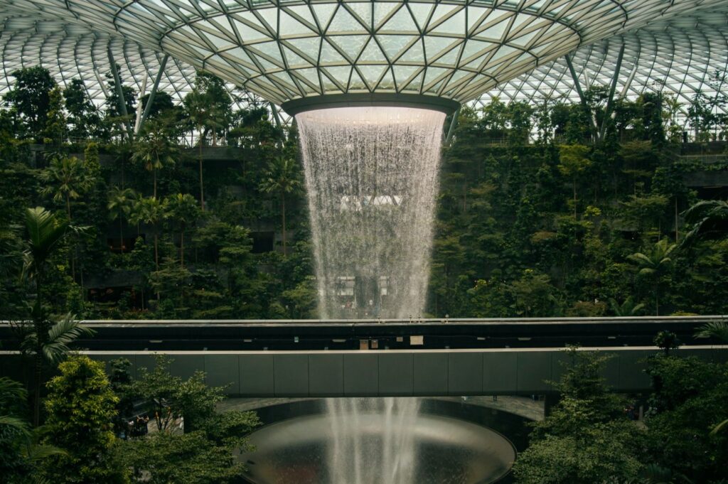 Indoor waterfall cascading from a glass dome roof, surrounded by lush green foliage. The scene conveys tranquility and a blend of nature and architecture.
