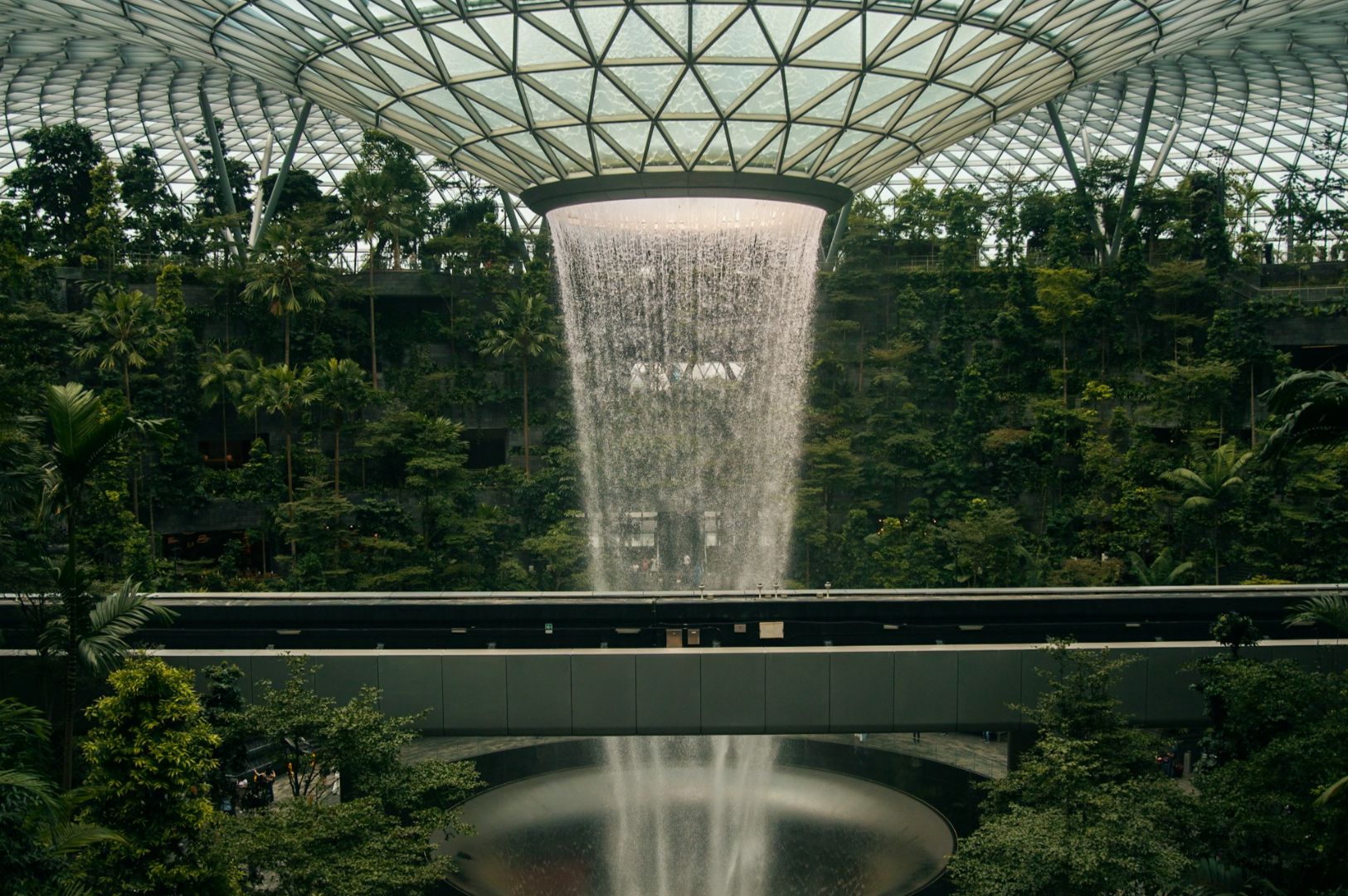 Indoor waterfall cascading from a glass dome roof, surrounded by lush green foliage. The scene conveys tranquility and a blend of nature and architecture.