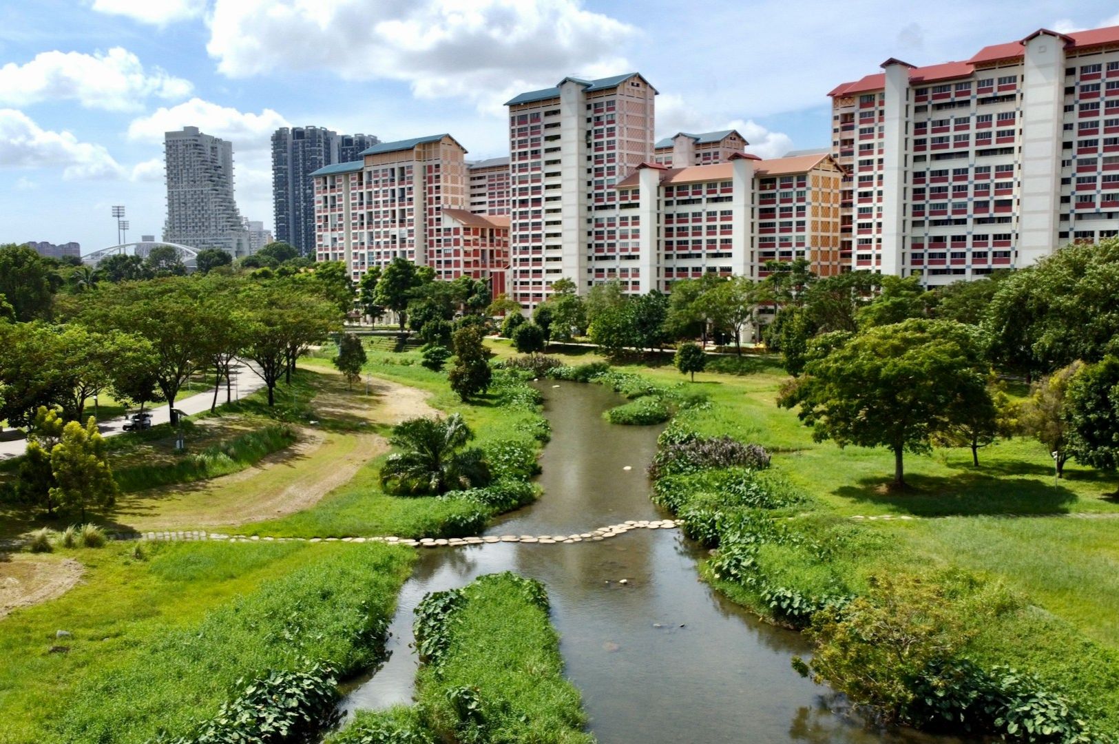 Urban park scene with a winding stream, lush green grass, and trees. Tall residential buildings tower in the background under a blue sky.