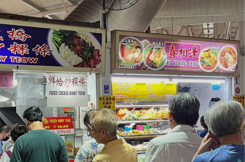 A busy indoor food court or hawker center with brightly lit stalls featuring signs for "Fried Cockle Kway Teow" and other dishes, with people standing in line.