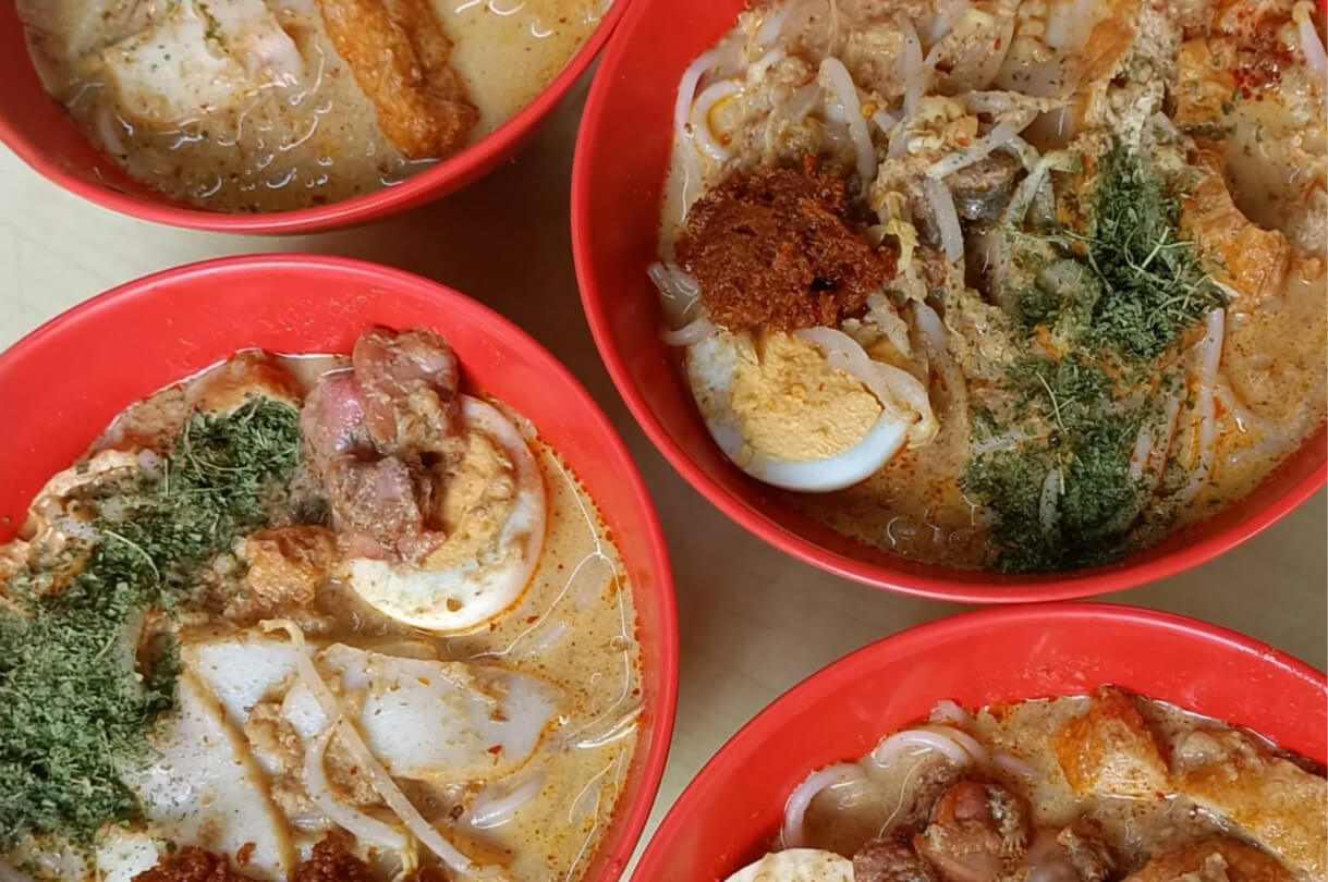 Four red bowls of Laksa featuring thick rice noodles in a creamy orange broth. The soup is topped with hard-boiled eggs, fish cakes, tofu puffs, a dollop of red sambal, and a generous sprinkle of chopped green laksa leaves.