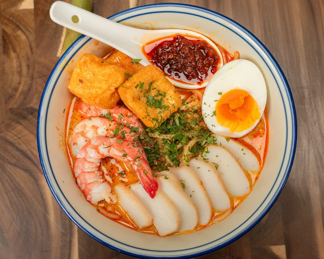 A comforting bowl of Nyonya laksa served in a ceramic bowl, filled with rich coconut-based broth, rice noodles, prawns, tofu puffs, and fresh herbs—an iconic Peranakan dish known for its fragrant spices and creamy texture