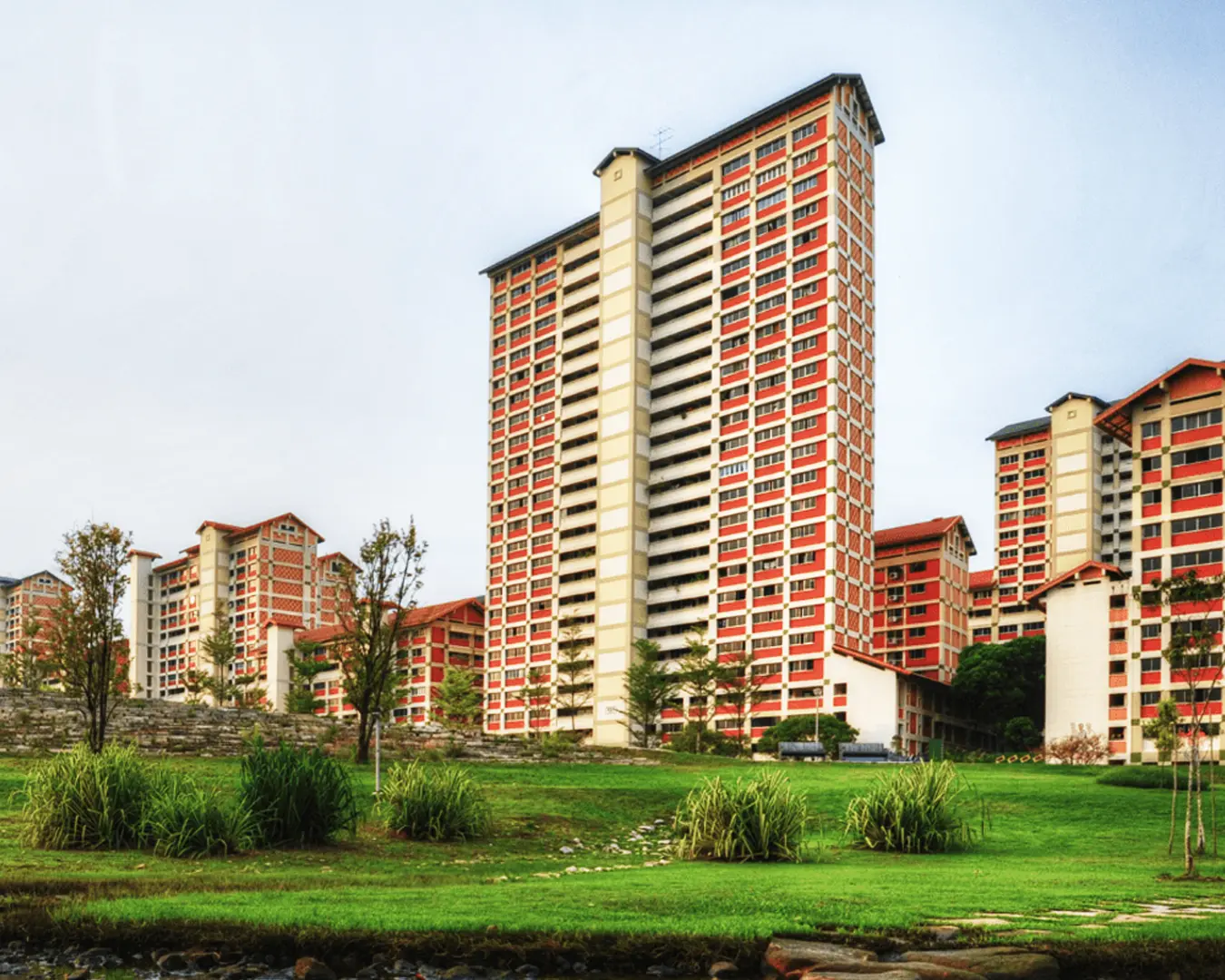 Tall, red and white apartment buildings stand prominently against a clear sky. Lush greenery and small trees surround the structures, creating a serene urban landscape.