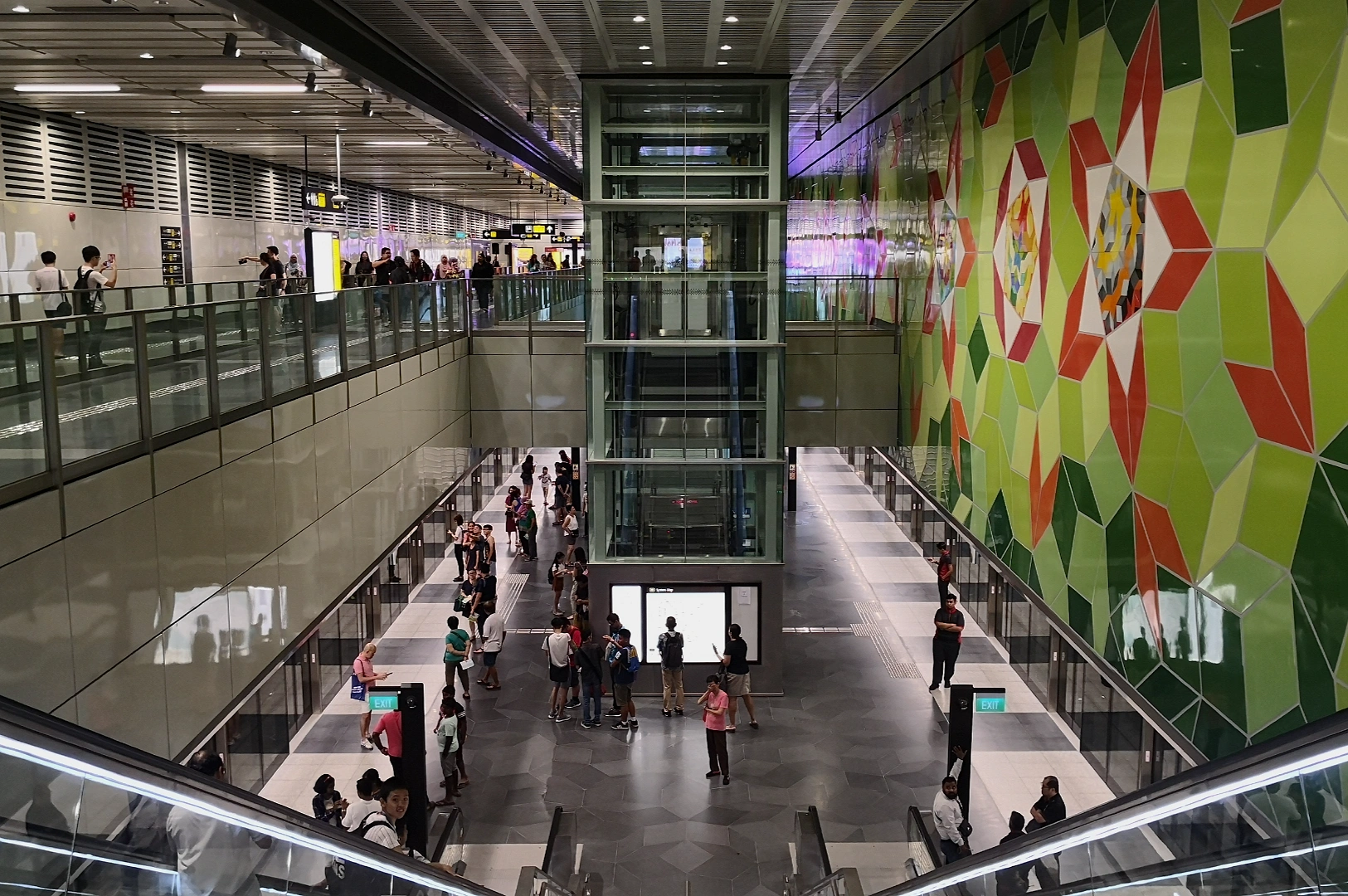“A busy subway station with people on two levels, featuring a vibrant, geometric green and red mural on the wall. The atmosphere is lively and bustling.”