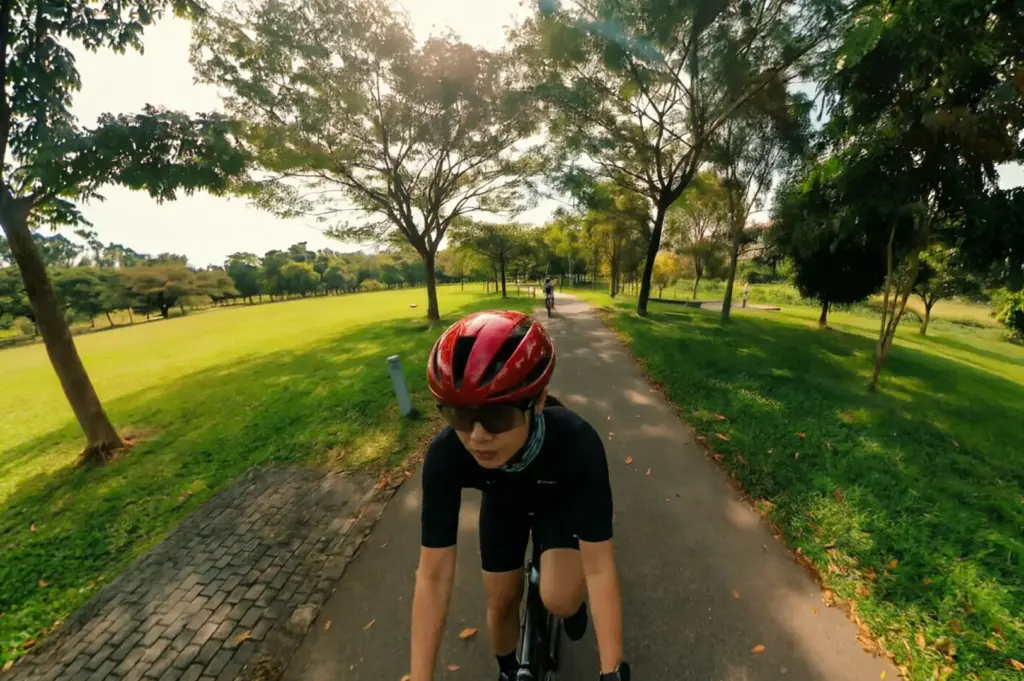 Cyclist wearing red helmet and black cycling gear riding along paved pathway through Bishan Park with expansive green lawns and mature trees creating shaded canopy on sunny day