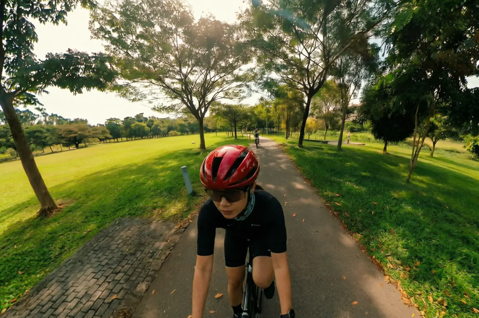 Cyclist wearing red helmet and black cycling gear riding along paved pathway through Bishan Park with expansive green lawns and mature trees creating shaded canopy on sunny day