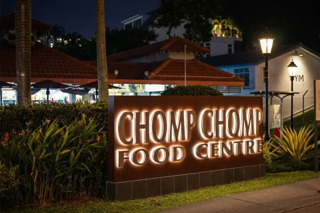 Illuminated Chomp Chomp Food Centre sign at night with distinctive red-tiled roof architecture and tropical landscaping in Serangoon Gardens