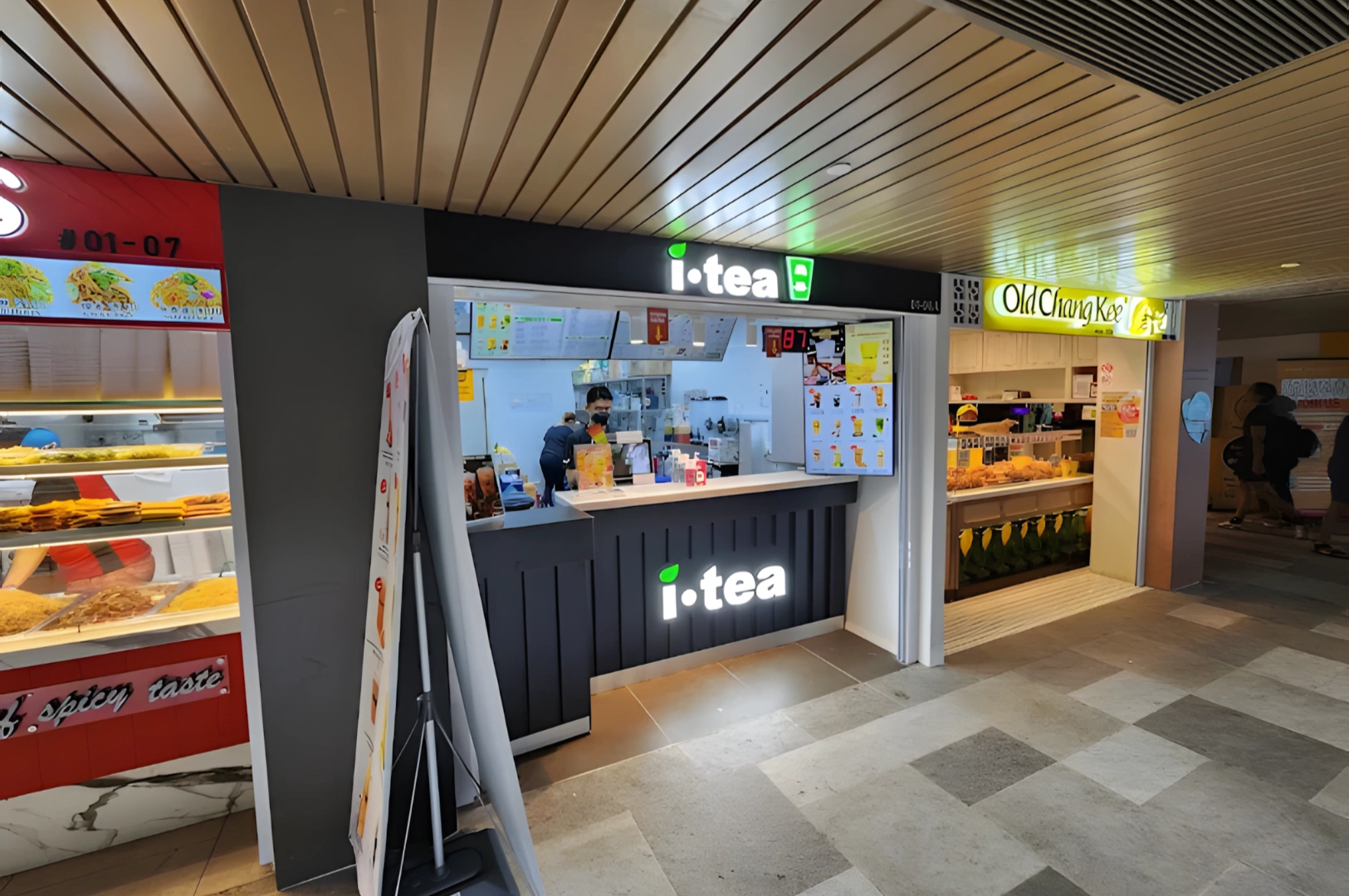 Two food stalls side by side in a mall, labeled "i-tea" and "Old Chang Kee." Customers are being served, creating a lively and inviting atmosphere