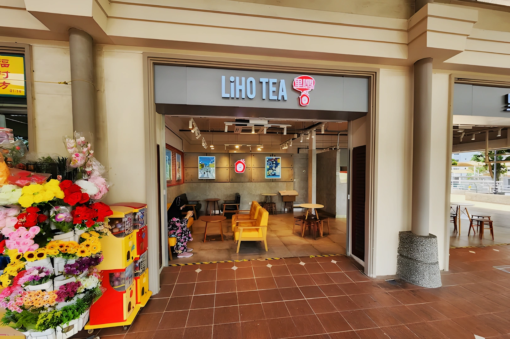 Entrance of a LiHO TEA store with a modern interior. Bright yellow seating inside contrasts with floral displays outside, creating an inviting atmosphere