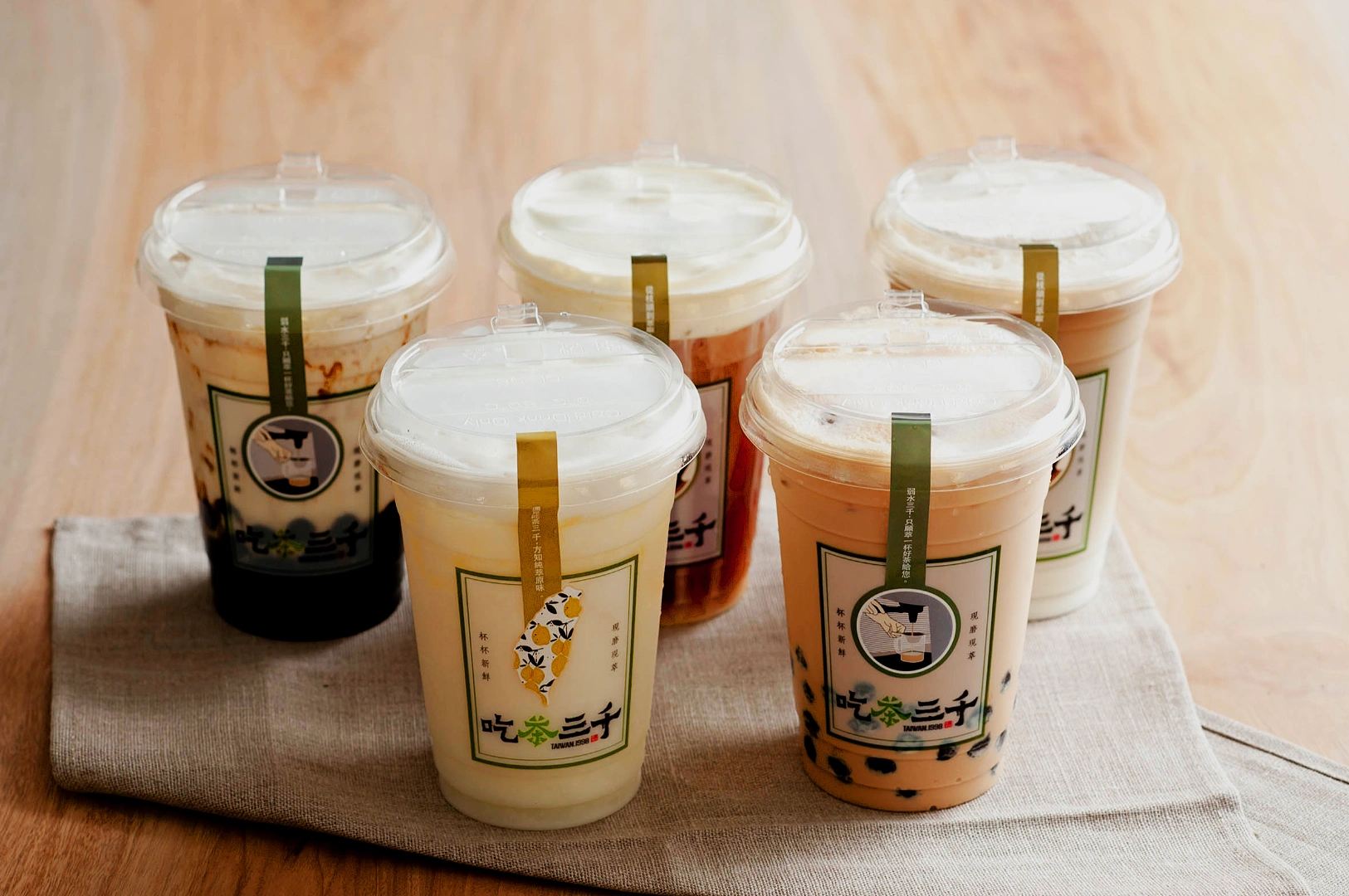 Five cups of bubble tea with varied toppings and creamy foam sit on a light-colored cloth atop a wooden surface, exuding a fresh and inviting vibe