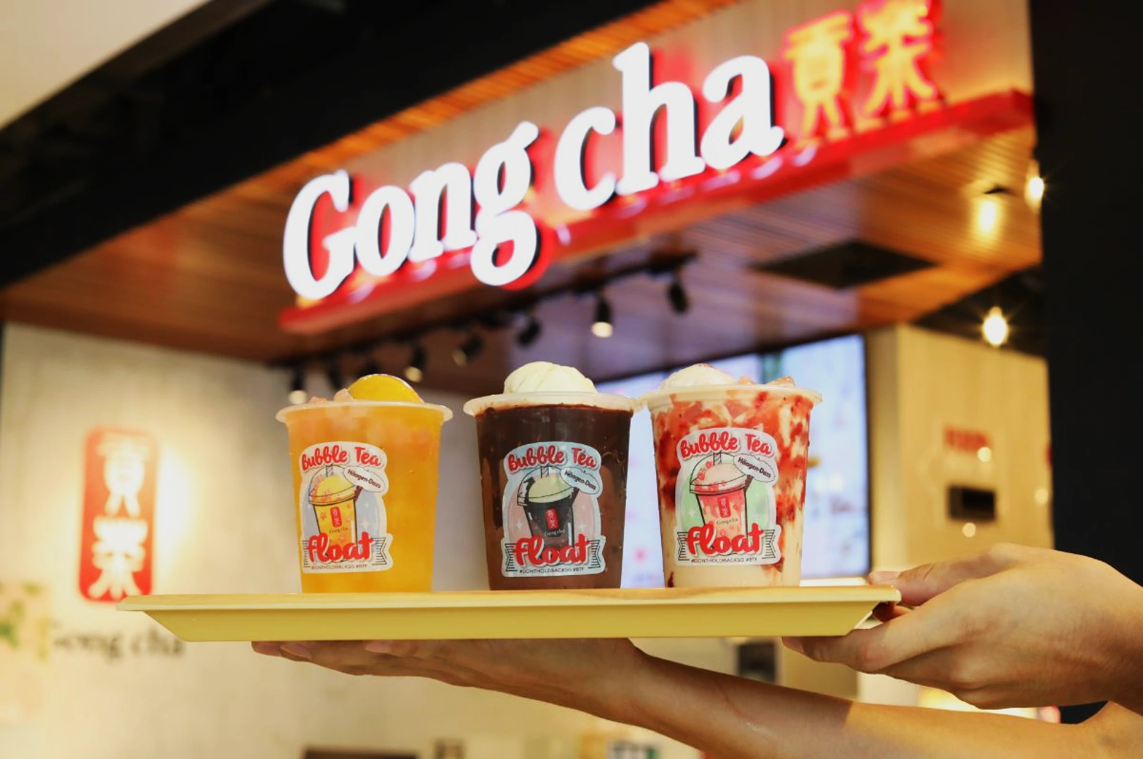 Three colorful bubble tea floats on a tray are held up in front of a Gong Cha store sign, creating a vibrant and refreshing display.