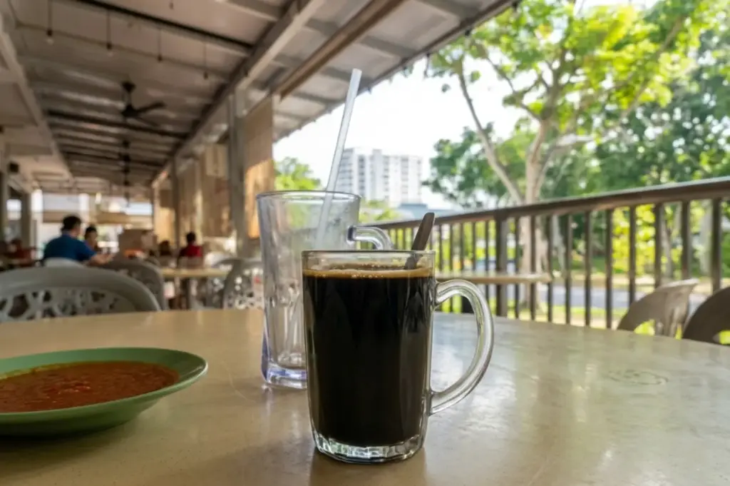 Black coffee in glass mug on table at open-air cafe with green sambal plate, trees and HDB buildings in Woodlands background