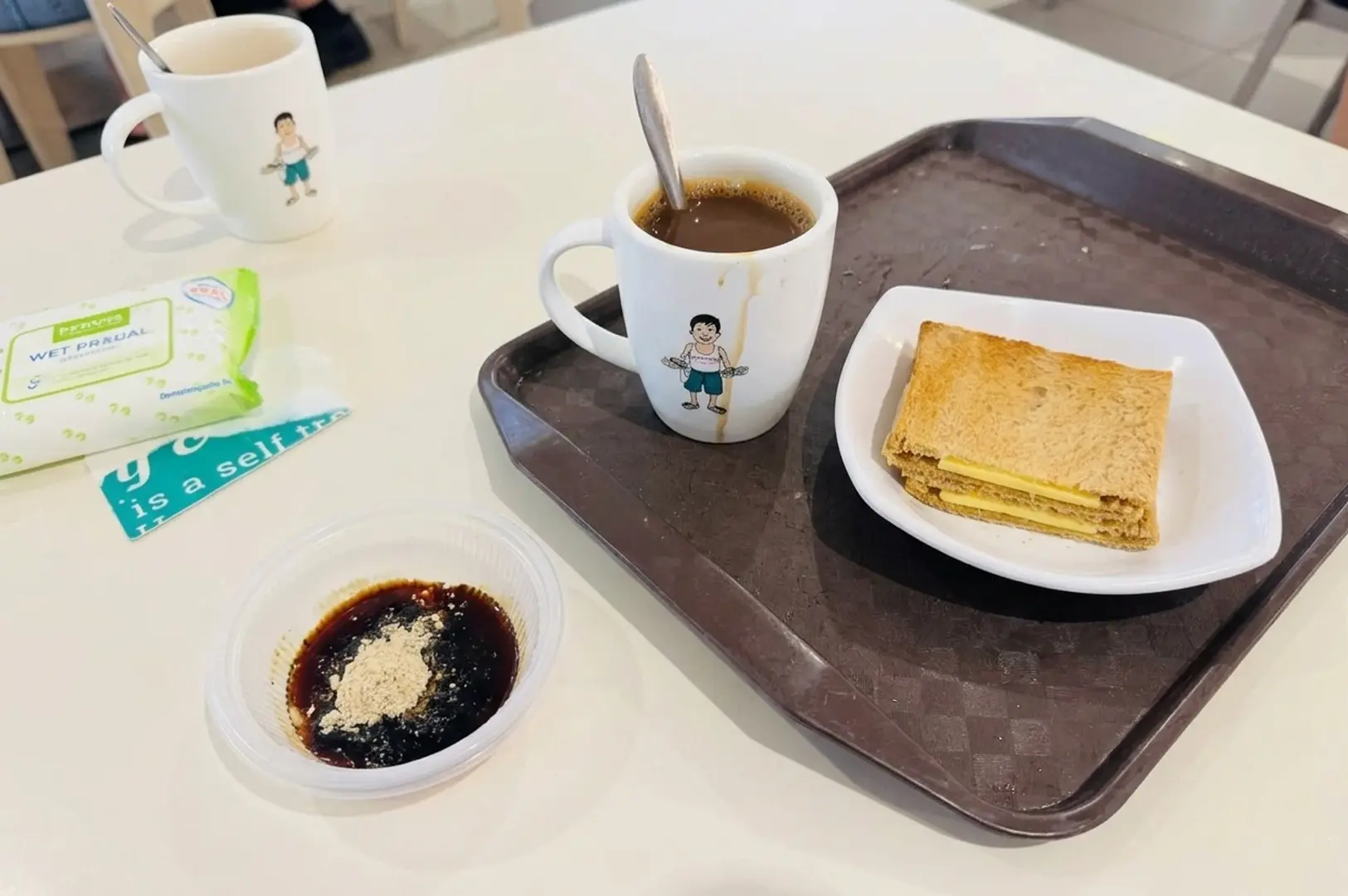Traditional Singaporean breakfast set with kopi coffee in character mug, kaya toast with butter, and condiments on brown tray