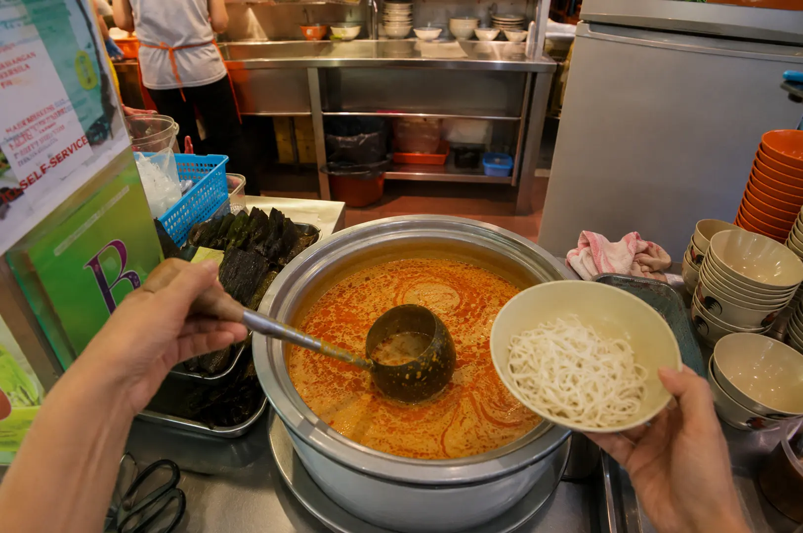 Hands scoop rich coconut laksa broth over fresh rice noodles at a laksa station in Katong, Singapore