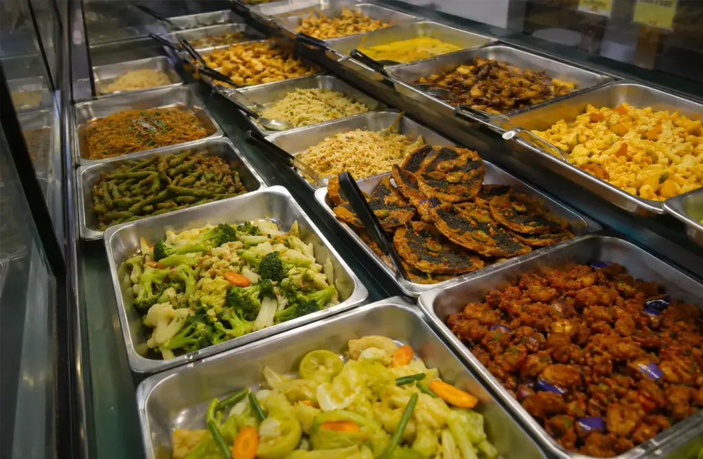 Display counter featuring various vegetarian dishes in metal trays including stir-fried vegetables, broccoli, cauliflower, and vegetarian patties