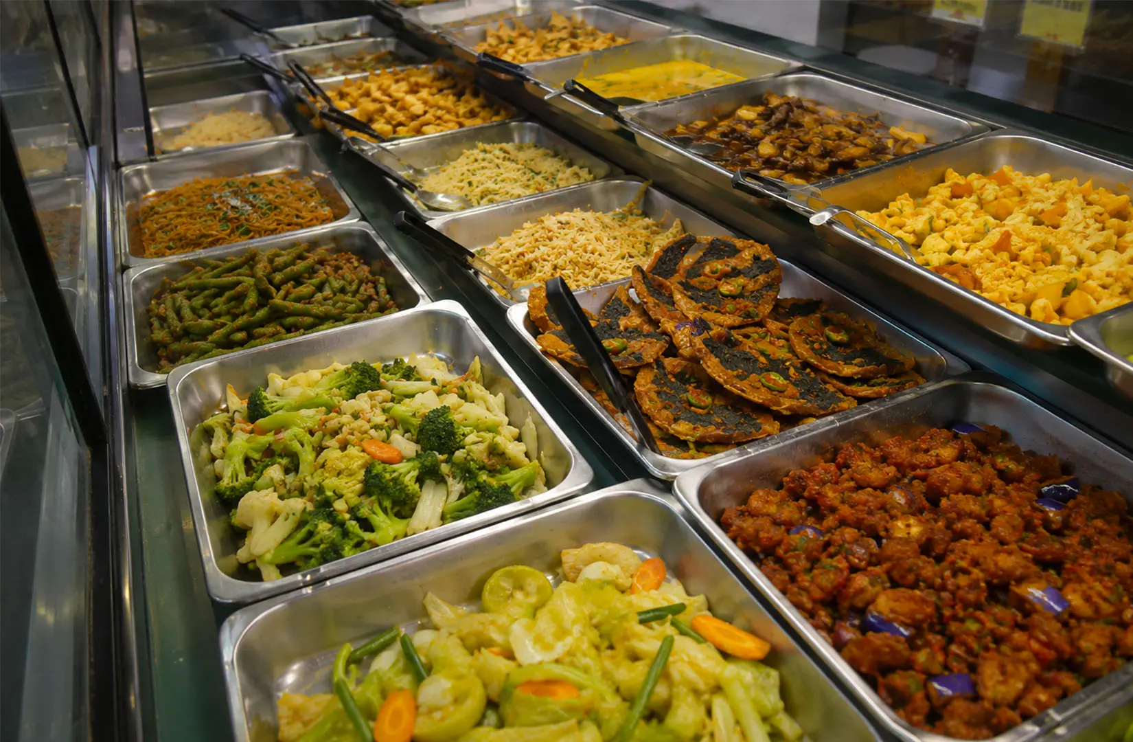 Display counter featuring various vegetarian dishes in metal trays including stir-fried vegetables, broccoli, cauliflower, and vegetarian patties