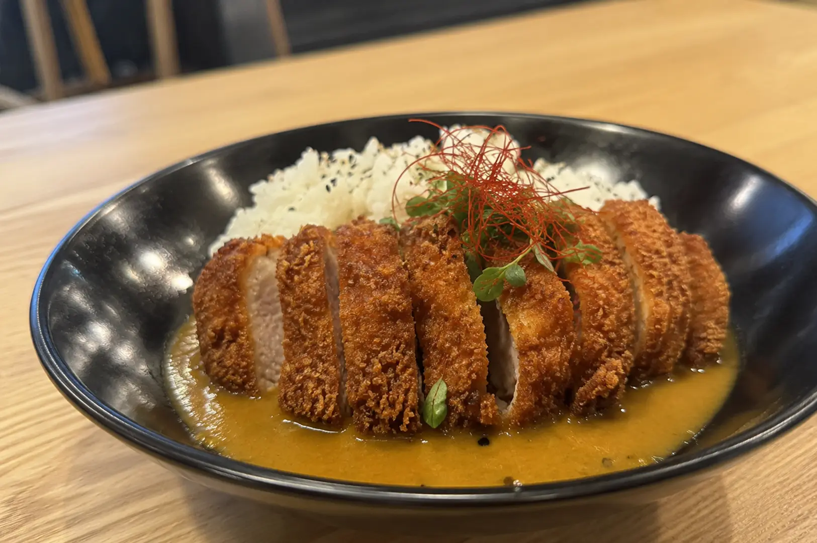 Close-up of Japanese katsu curry featuring sliced breaded pork cutlet over rice with rich curry sauce, garnished with red threads and microgreens in a black bowl