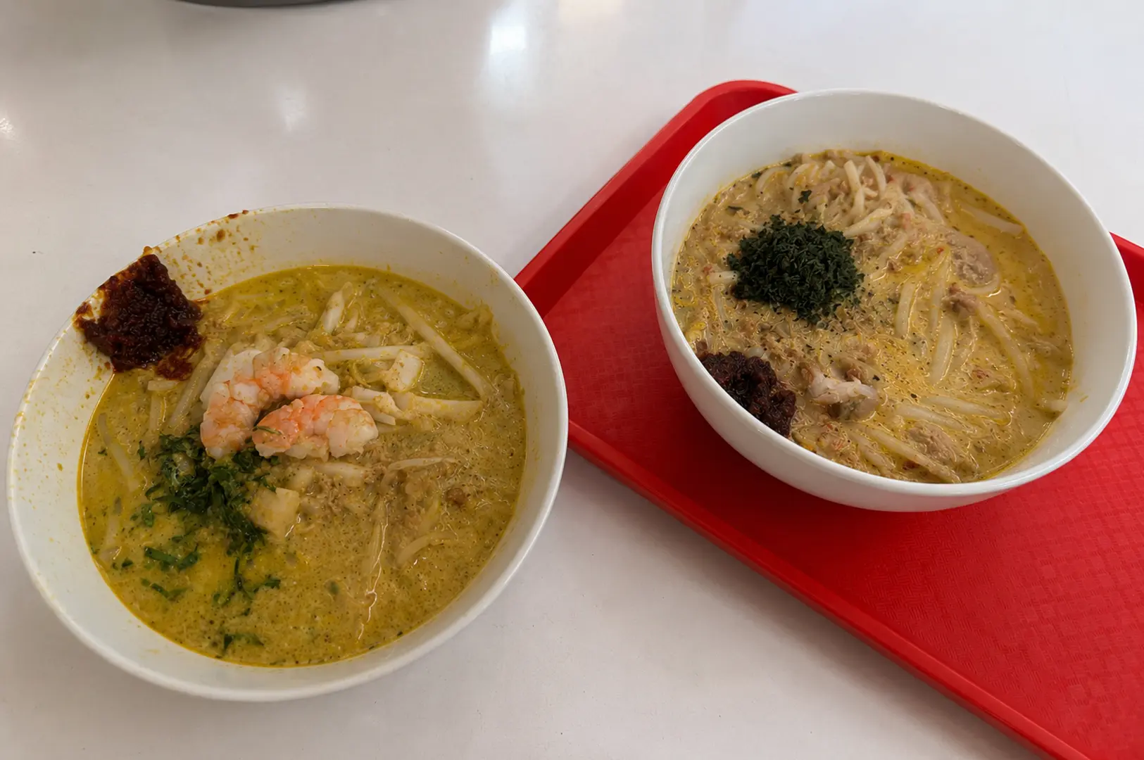 Two bowls of Katong laksa served side by side topped with prawns, seafood, herbs, sambal, and creamy coconut curry broth with rice noodles