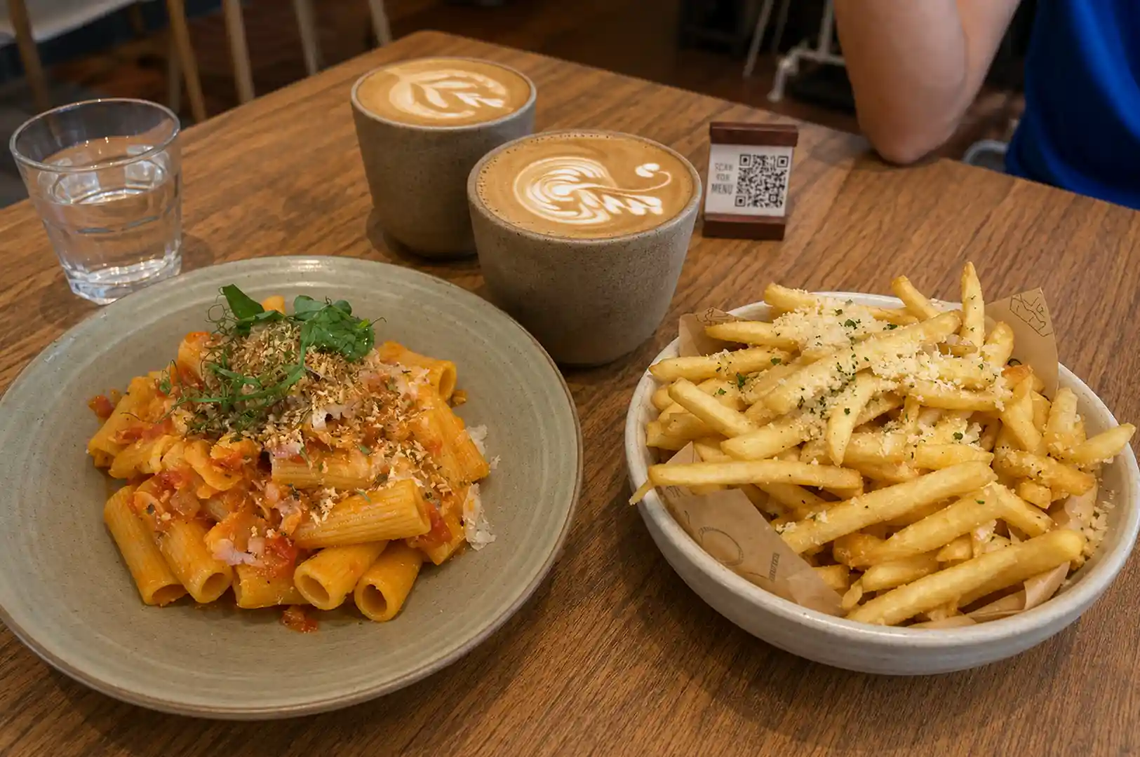 Table setting featuring two lattes with latte art, a plate of penne pasta, and a bowl of french fries at a cafe