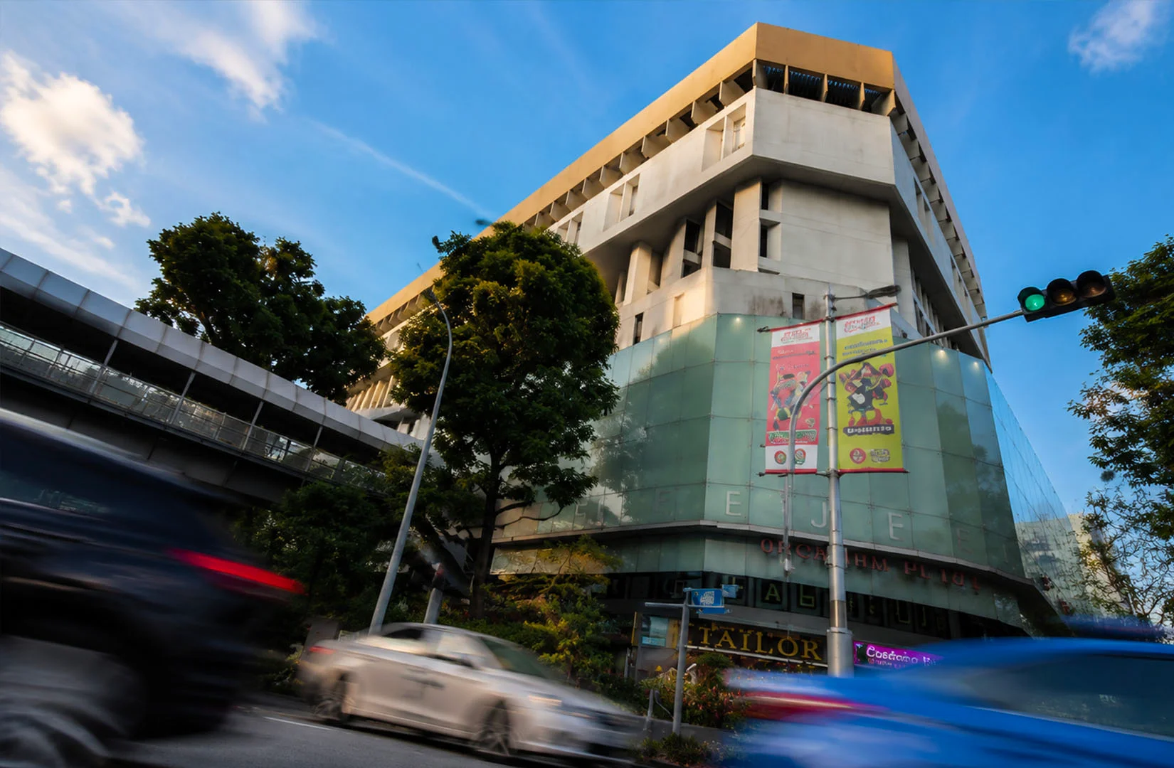 Exterior view of Orchard Plaza along Orchard Road, showing its aging façade, surrounding trees, and busy street traffic that reflects the building’s everyday urban presence.