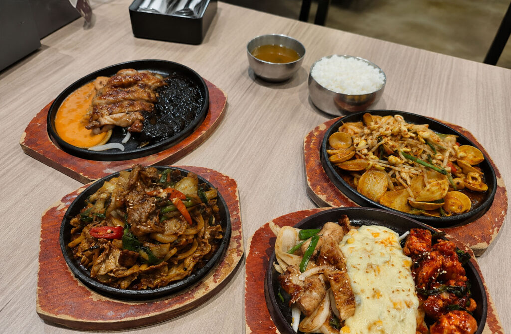A table filled with Korean dishes served on sizzling hot plates, featuring grilled meats and savoury sides in a casual dining setting.